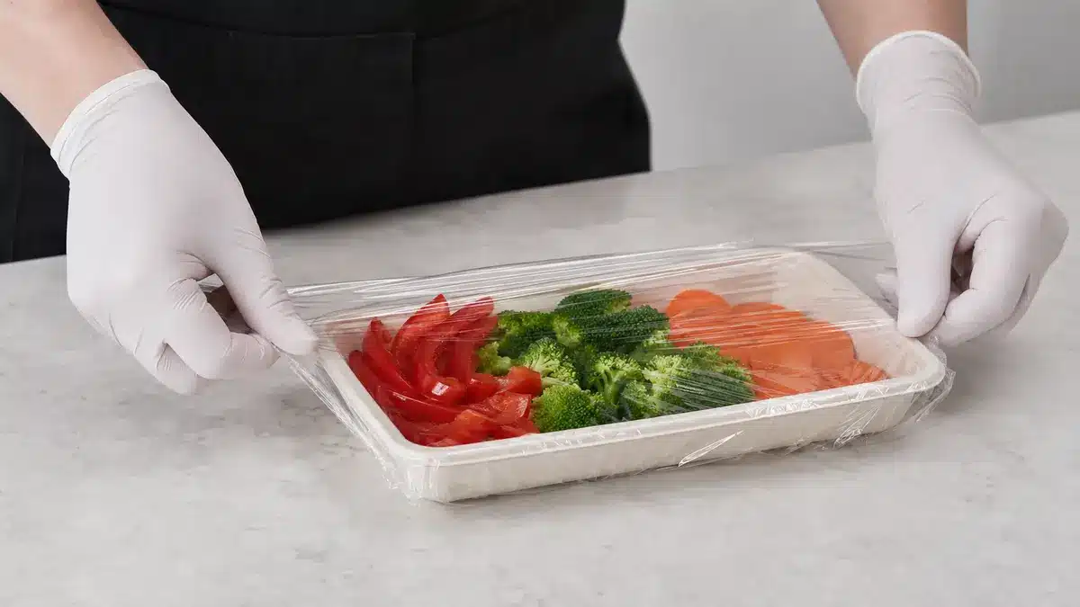 Gloved hands wrapping a shallow fresh-produce tray with transparent cling film